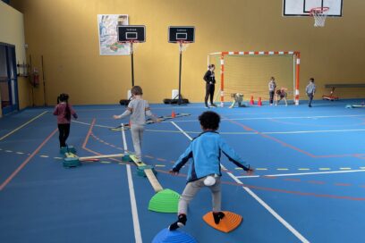 Photo d'un parcours sportif pour enfants dans un gymnase, on voit un petit marcher sur des marques et deux autres marcher sur des mini poutres en bois à ras du sol - Agrandir l'image, fenêtre modale