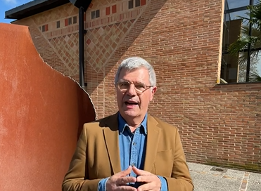 Le Maire Dominique Fouchier parle face à la caméra, en extérieur, devant un bâtiment aux briques rouge clair et une œuvre d'art métallique - Agrandir l'image, fenêtre modale