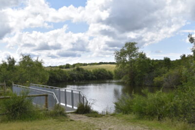 Photo d'un paysage naturel, journée ensoleillée avec quelques nuages, on voit un plan d'eau et une passerelle qui passe par-dessus - Agrandir l'image 1 sur 3, fenêtre modale