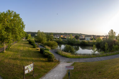 Photo panorama du parc de la Paderne, chemin piétonnier qui se sépare à gauche et droite et derrière, un cours d'eau. On aperçoit des habitations à l'arrière-plan