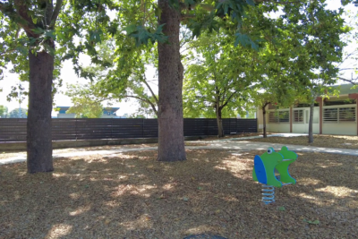 Photo d'une cour d'école entièrement ombragée grâce à de grands arbres, on voit un jeu à bascule pour enfant et le sol est en copeaux de bois - Agrandir l'image 2 sur 3, fenêtre modale