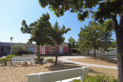 Photo de la cour d'école, au premier plan un banc blanc en pierre, derrière, un sol en copeaux de bois, des arbres qui permettent d'avoir de l'ombre - Agrandir l'image 1 sur 3, fenêtre modale