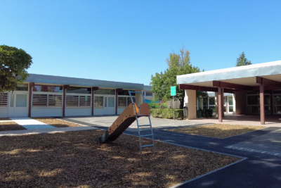 Photo d'une cour d'école ombragée, dont le sol est fait de copeaux de bois. On voit un toboggan, un préau. - Agrandir l'image 1 sur 3, fenêtre modale