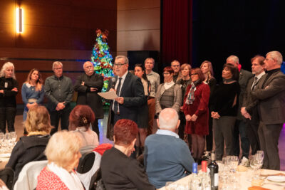 Photo du Maire Frédéric Parre qui fait un discours, on voit derrière lui les élus de la majorité rassemblés, et dans l'arrière-plan un sapin de Noël décoré et lumineux. Au premier plan, des séniors de dos, tournés vers le Maire pour l'écouter - Agrandir l'image 8 sur 14, fenêtre modale