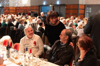 Photo de l'élue Rachida Lucazeau qui salue deux séniors à table. Derrière eux on voit les autres tables avec les nombreux séniors rassemblés pour manger - Agrandir l'image 9 sur 14, fenêtre modale