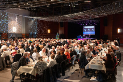 Photo d'ensemble de la salle, on voit les rangées de table alignées et les très nombreux séniors attablés. Au-dessus, des guirlandes suspendues, et sur le mur du côté gauche de la salle, des projections lumineuses comme de flocons de neige - Agrandir l'image 11 sur 14, fenêtre modale