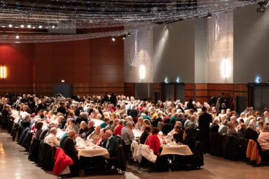 Photo globale de la salle où sont installées de longues tables, autour desquelles des centaines de personnes âgées sont assises à discuter et manger. Au plafond, il y a des guirlandes lumineuses pour déco.