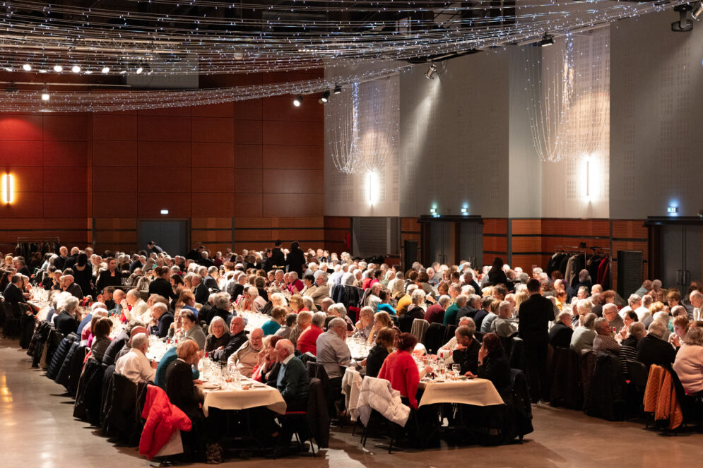 Photo globale de la salle où sont installées de longues tables, autour desquelles des centaines de personnes âgées sont assises à discuter et manger. Au plafond, il y a des guirlandes lumineuses pour déco.