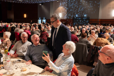 Le Maire Frédéric Parre échange avec des séniors qui rient, une dame montre au Maire quelque chose sur ton téléphone. Derrière eux, on voit les autres tables alignées avec de très nombreux séniors rassemblés - Agrandir l'image 1 sur 14, fenêtre modale