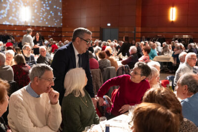 Photo du Maire qui échange avec des personnes âgées assises à table, dans l'arrière-plan, on voit de nombreuses personnes assises également à discuter - Agrandir l'image 4 sur 14, fenêtre modale