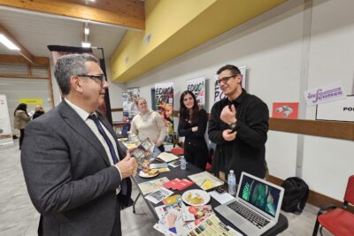Le Maire échange avec un homme debout derrière un stand sur lequel il y a des documents, des carambars à disposition, et derrière sur le mur, une affichette "info jeunes" - Agrandir l'image 11 sur 13, fenêtre modale