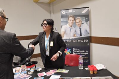 Le Maire serre la main d'une femme en tailleur, avec lunettes, debout à un stand sur lequel on a de la documentation posée. Derrière le stand, on peut voir un kakémono avec écrit "votre story se construit avec eux" et "cité des formations & des apprentissages" - Agrandir l'image 13 sur 13, fenêtre modale