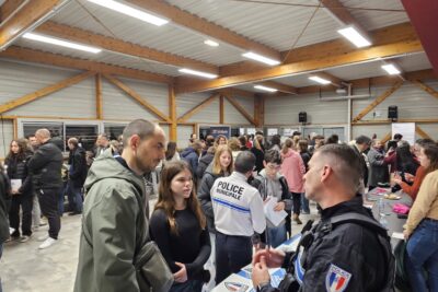 Une jeune fille et un jeune homme écoutent un policier municipal au stand d'information de la police municipale. On voit également dans la salle d'autres jeunes rassemblés autour d'autres stands d'information - Agrandir l'image 3 sur 13, fenêtre modale
