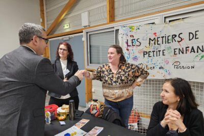 Le Maire serre la main d'une femme qui se tient debout à un stand, derrière lequel une grille est debout avec une sorte de banderole sur laquelle il est écrit "les métiers de la petite enfance, métier de passion" - Agrandir l'image 7 sur 13, fenêtre modale