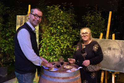 Un homme et une femme sourient à l'objectif, ils mangent, debout à côté d'un tonneau où sont posées leurs petites assiettes - Agrandir l'image 9 sur 22, fenêtre modale