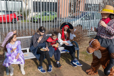 Une famille mange le goûter dans le parc, assise sur un banc, il y a une mère en pirate qui sourit à l'objectif, et les enfants à côté en princesse et en super-héros - Agrandir l'image 12 sur 18, fenêtre modale