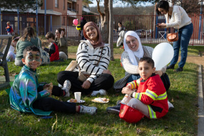 Une famille en train de prendre le goûter par terre, deux femmes tout sourire et deux jeunes garçons, un déguisé en pompier et l'autre avec une cape colorée et brillante - Agrandir l'image 13 sur 18, fenêtre modale