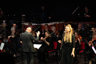 Plan d'ensemble orchetsre en fon de scène directeur musical dos au public debout en costume noir élégant, la choriste cheveux retenus par in chignon sur le sommet du crâne, debout en combinaison noire fluide, chante au micro face public à droite de l'image - Agrandir l'image 2 sur 8, fenêtre modale