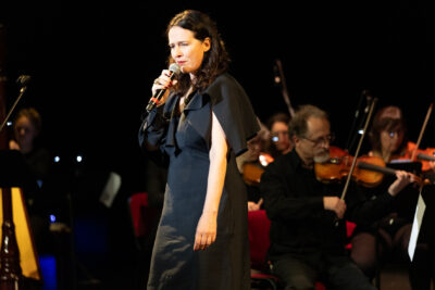 Claire Diterzi debout micro contre sa bouche dans une robe longue de soirée noire, le premier violon, apparaît à sa gauche - Agrandir l'image 6 sur 8, fenêtre modale