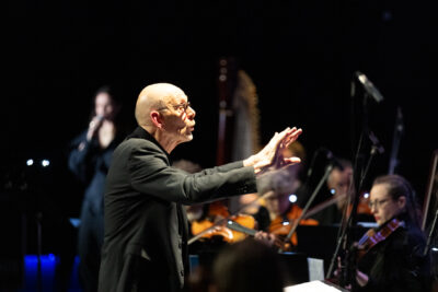 Le chef d'porechter debout de profil vers la droite de l'image dirige ses musiciens de manière expressive main tendue à hauteur de visage - Agrandir l'image 8 sur 8, fenêtre modale