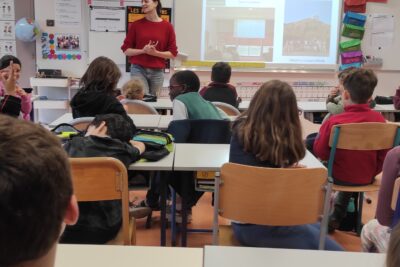 Photo d'une salle de classe d'école primaire, prise du fond de la classe, on voit une quinzaine d'enfants assis qui écoutent une jeune femme avec un pull rouge leur expliquer quelque chose, pendant qu'un diaporama est projeté sur lequel il est écrit "une semaine dans la vie de chercheuse" - Agrandir l'image 2 sur 6, fenêtre modale