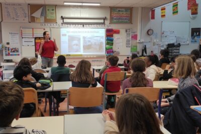 Photo d'une salle de classe d'école primaire, prise du fond de la classe, on voit une quinzaine d'enfants assis qui écoutent une jeune femme avec un pull rouge leur expliquer quelque chose, pendant qu'un diaporama est projeté sur lequel il est écrit "une semaine dans la vie de chercheuse" - Agrandir l'image 3 sur 6, fenêtre modale