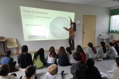 Photo dans une salle, des jeunes écoliers sont assis par terre et écoutent une jeune femme présenter un diaporama autour de son stage de recherche - Agrandir l'image 5 sur 6, fenêtre modale