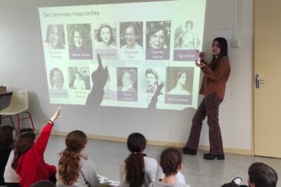Photo dans une salle, des jeunes écoliers sont assis par terre et écoutent une jeune femme présenter un diaporama sur lequel on voit des portraits de femmes scientifiques. Certains enfants lèvent la main - Agrandir l'image 6 sur 6, fenêtre modale