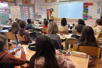 Photo d'une salle de classe d'école primaire, prise du fond de la classe, on voit une quinzaine d'enfants assis qui écoutent une jeune femme avec un pull rouge leur expliquer quelque chose, pendant qu'un diaporama est projeté sur lequel il est écrit "une semaine dans la vie de chercheuse" - Agrandir l'image 1 sur 6, fenêtre modale