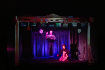 Décor de scène dans le noir où les personnages des deux comédiennes vêtues de tuniques traditionnelles japonaises se tiennent sous un auvent décoré finement dans un esprit japonisant, où les lumières roses, bleues et violette dominent. - Agrandir l'image 6 sur 7, fenêtre modale