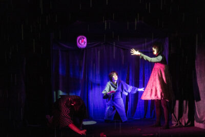 Dans le théâtre japonais qui tient de lieu de départ et d'arrivée de la pièce, le personnage principal se tient à présent debout de profil perchée au-dessus des deux autres personnages en position de soumission ou de crainte/respect, elle ouvre grand sa main illuminée par une lumière éclatante qui tranche avec les couleurs roses-violette du reste de la scène - Agrandir l'image 7 sur 7, fenêtre modale