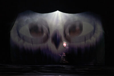 Debout de profil en fond de scène un personnage de profil tenant une loupiote au-dessus de sa tête, en manteau, se confronte à l'image effrayante d'une tête de hibou géant nébuleuse qui occupe tout le fond de l'espace scénique - Agrandir l'image 2 sur 7, fenêtre modale