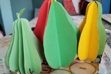 photo de travail plastique réalisé en atelier à base de carton découpé et coloré pour former une nature morte en 3 dimensions représentants des fruits en forme de poires rassemblés dans un même espace et dont les couleurs associent des lames rouges, jaunes et vertes qui figurent les teintes du fruit dans sa version naturelle