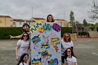 Photo de 6 femmes rassemblées dans une cour d'école autour d'un panneau bleu sur lequel il est écrit "journée de la super atsem" et des illustrations d'elles façon super-héroïnes - Agrandir l'image 4 sur 8, fenêtre modale