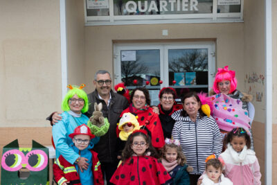 Photo portrait de groupe devant l'entrée de la maison de quartier Pahin, 6 enfants déguisés et derrière eux les adultes dont Monsieur le Maire Frédéric Parre - Agrandir l'image 1 sur 28, fenêtre modale