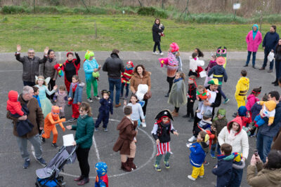 Photo en plongée d'une quinzaine de familles rassemblées sur un terrain de foot en béton, les enfants sont déguisés, on voit le Maire qui fait signe à l'objectif en souriant - Agrandir l'image 2 sur 28, fenêtre modale