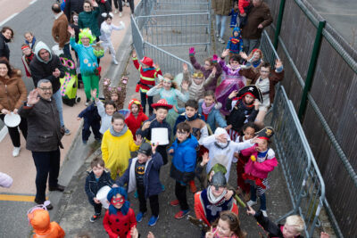 Photo du défilé de carnaval, en plongée, mené par une vingtaine d'enfants déguisés qui lèvent les bras pour faire coucou à l'objectif - Agrandir l'image 7 sur 28, fenêtre modale