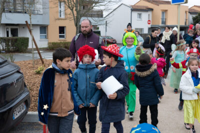 Photo du défilé avec trois garçons au premier plan qui portent une écharpe de magicien, une perruque de clown rouge et un chapeau rouge respectivement, et celui sur la droite a un seau sur lequel il abat une cuillère pour faire du bruit, derrière eux on voit le défilé qui suit avec d'autres enfants plus petits déguisés accompagnés de leurs parents - Agrandir l'image 26 sur 28, fenêtre modale