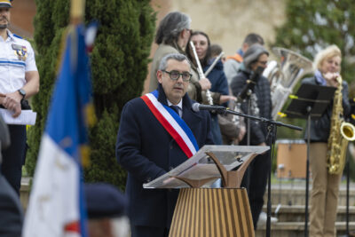 Photo du Maire qui fait un discours. Derrière lui, on voit un homme de la police nationale en uniforme, et de l'autre côté également derrière lui, l'orchestre - Agrandir l'image 9 sur 16, fenêtre modale