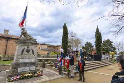 Photo du Monument aux morts avec à côté les porteurs de drapeaux, devant le monument, les gerbes de fleurs. Derrière les porteurs de drapeau, on voit l'orchestre également. - Agrandir l'image 10 sur 16, fenêtre modale