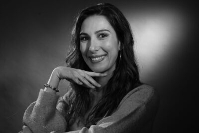 Photo d'une femme assise sur une chaise en osier, le dossier devant, elle appuie son bras gauche et a sa main droite sous le menton, elle a les cheveux longs et sourit à l'objectif - Agrandir l'image 3 sur 21, fenêtre modale