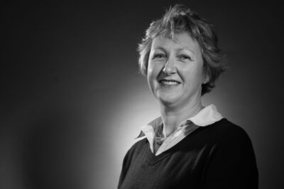 Photo portrait en noir et blanc d'une femme assise, les mains posées à plat sur les jambes, elle a les cheveux courts et un pull avec un col de chemise claire, elle sourit à l'objectif - Agrandir l'image 4 sur 21, fenêtre modale