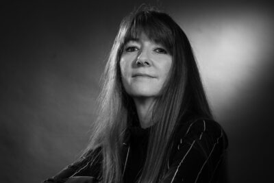 Photo portrait en noir et blanc d'une femme assise sur une chaise en osier, le dossier face à elle, elle s'appuie les bras croisés dessus - Agrandir l'image 12 sur 21, fenêtre modale