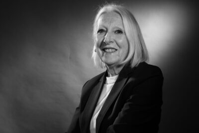 Photo portrait en noir et blanc d'une femme assise, de biais, les jambes croisées, avec un haut de tailleur. Elle sourit à l'objectif - Agrandir l'image 21 sur 21, fenêtre modale