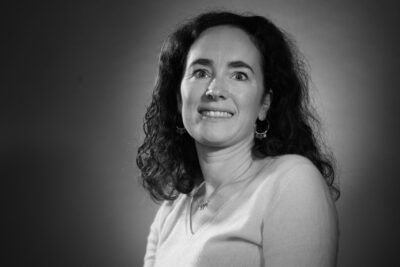 Photo portrait en noir et blanc d'une femme assise avec un haut clair, les cheveux un peu en-dessous des épaules, la silhouette de biais et le visage de face, elle sourit à l'objectif - Agrandir l'image 1 sur 21, fenêtre modale