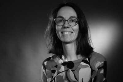 Photo portrait en noir et blanc d'une femme aux cheveux qui tombent à l'épaule, avec des lunettes, elle croise les mains et sourit - Agrandir l'image 7 sur 21, fenêtre modale