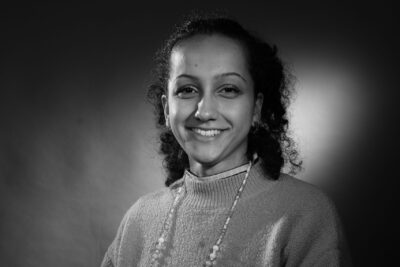 Photo d'une femme en noir et blanc, souriante, elle a des lunettes autour du cou, la main gauche sur sa jambe croisée par-dessus la droite - Agrandir l'image 18 sur 21, fenêtre modale