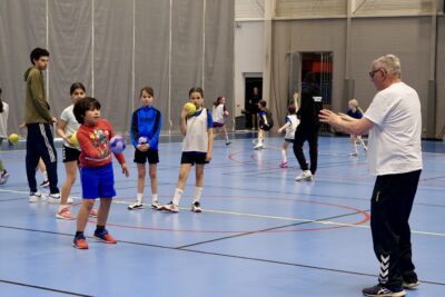 Photo d'un enfant qui envoie la balle à un adulte sur le terrain de handball, d'autres enfants derrière - Agrandir l'image 10 sur 14, fenêtre modale