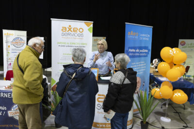 Une femme derrière le stand Axeo Services échange avec trois personnes âgées - Agrandir l'image 3 sur 20, fenêtre modale