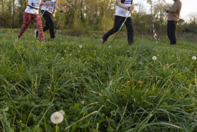Photo de participantes à la marche nordique qui passent à côté d'un pêcheur, dans un chemin d'herbe, la photo est prise en contreplongée avec vue au premier plan sur un pissenlit - Agrandir l'image 5 sur 32, fenêtre modale